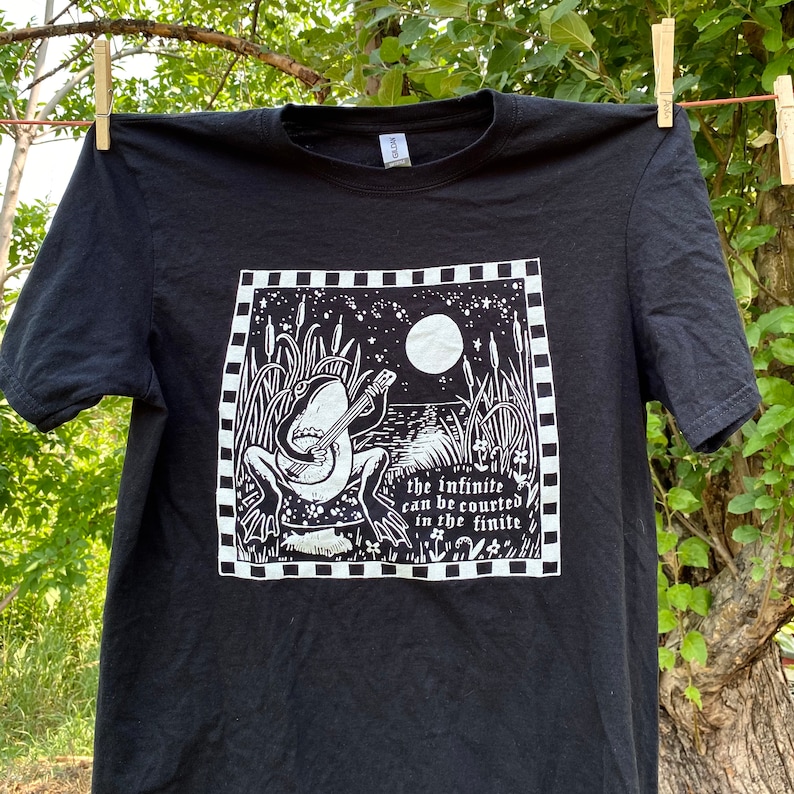 May include: Black t-shirt featuring a white graphic print of a frog playing a banjo under a full moon. The text "the infinite can be courted in the finite" is printed below the frog.