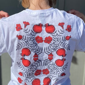 May include: White t-shirt with a red and black pomegranate design. The design features pomegranates, leaves, and flowers.