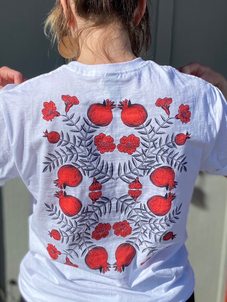 May include: White t-shirt with a red and black pomegranate design. The design features pomegranates, leaves, and flowers.