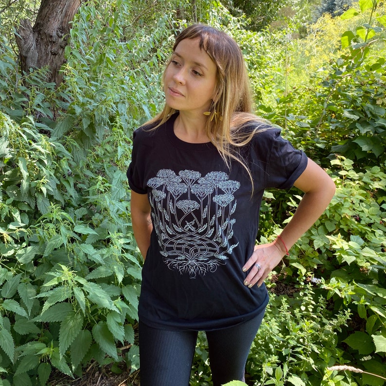 K&ouml;nnte beinhalten: Ein schwarzes T-Shirt mit einem wei&szlig;en Aufdruck einer Gruppe von Schafgarbenbl&uuml;ten. Der Aufdruck ist detailliert und kunstvoll und zeigt die St&auml;ngel, Bl&auml;tter und Bl&uuml;ten der Pflanze.