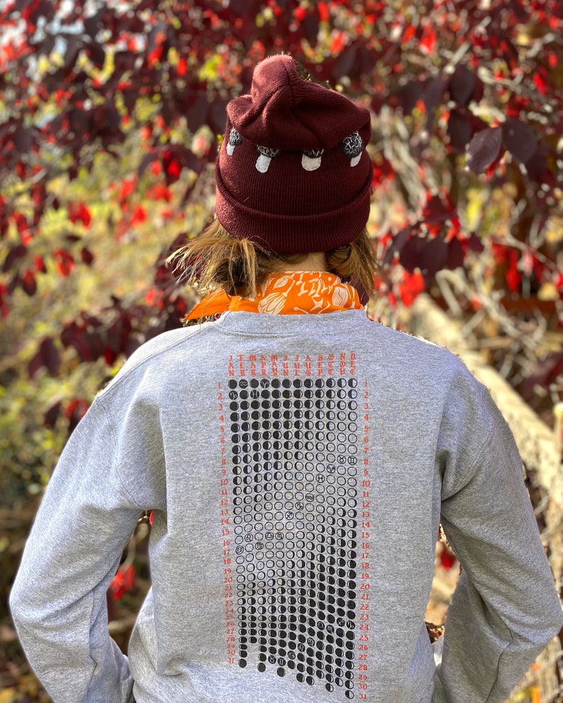 K&ouml;nnte beinhalten: Ein graues Sweatshirt mit einem schwarz-wei&szlig;en Mondphasenkalender, der auf der R&uuml;ckseite aufgedruckt ist. Der Kalender zeigt die Mondphasen f&uuml;r jeden Tag des Monats. Das Sweatshirt hat eine kastanienbraune M&uuml;tze mit wei&szlig;en Pilz-Akzenten oben.