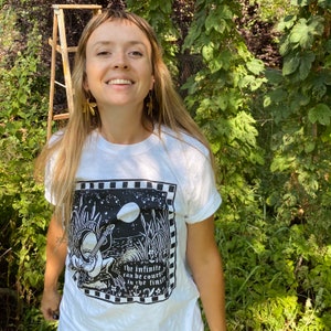 May include: A white t-shirt with a black and white illustration of a person playing a musical instrument. The text on the shirt reads "the infinite can be count in the finite".