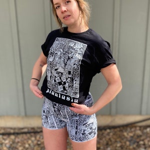 May include: A black t-shirt with a white graphic print of a plant-themed design. The text "plantasia" is visible on the shirt. The person is wearing black and white patterned shorts.