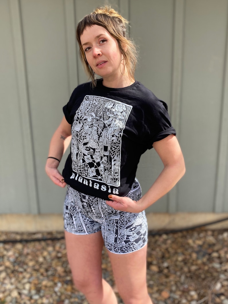 May include: A black t-shirt with a white graphic print of a plant-themed design. The text "plantasia" is visible on the shirt. The person is wearing black and white patterned shorts.