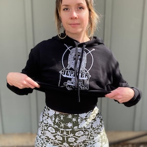 May include: A black hoodie with a white graphic of a person with a beard and the text "Jupiter is Home". The person is wearing a crown and is surrounded by a circle. The hoodie is being held up by a person wearing green and white floral print shorts.
