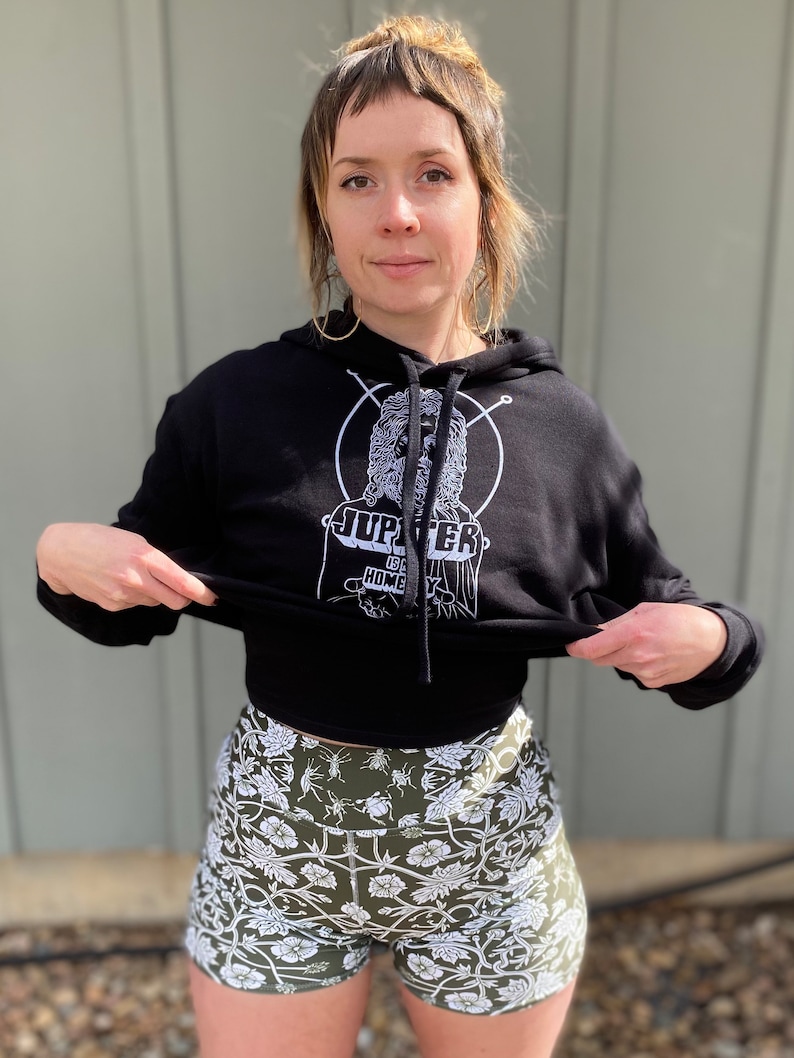 May include: A black hoodie with a white graphic of a person with a beard and the text "Jupiter is Home". The person is wearing a crown and is surrounded by a circle. The hoodie is being held up by a person wearing green and white floral print shorts.
