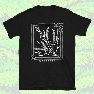 May include: Black t-shirt featuring a white illustration of lavender sprigs within a square frame. The design includes astrological symbols and the word "Lavender" in a stylized font. The shirt is made of a soft material.