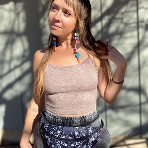 May include: A woman wearing a brown tank top and black pants with a black and white fanny pack with a psychedelic design. She is wearing colorful beaded earrings.