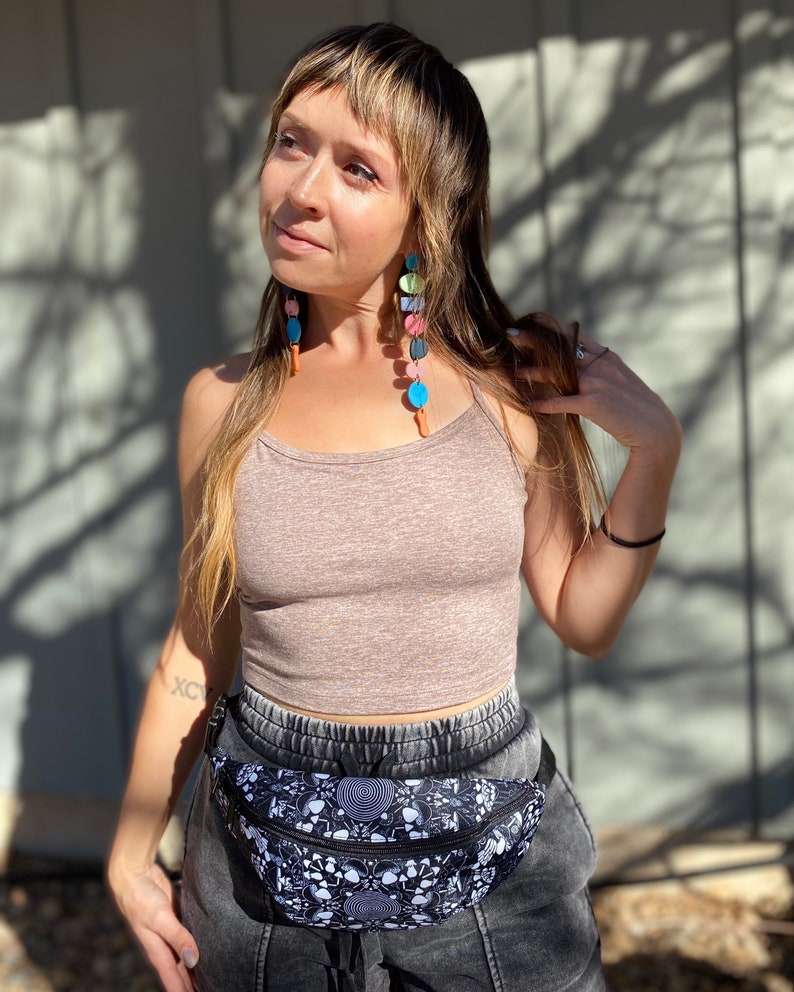 May include: A woman wearing a brown tank top and black pants with a black and white fanny pack with a psychedelic design. She is wearing colorful beaded earrings.