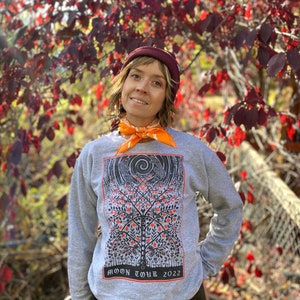 K&ouml;nnte beinhalten: Ein graues Sweatshirt mit einem schwarzen und orangenen Aufdruck eines Baumes mit V&ouml;geln und dem Text "MOON TOUR 2022". Die Person, die das Sweatshirt tr&auml;gt, steht vor einem Busch mit roten Bl&auml;ttern.