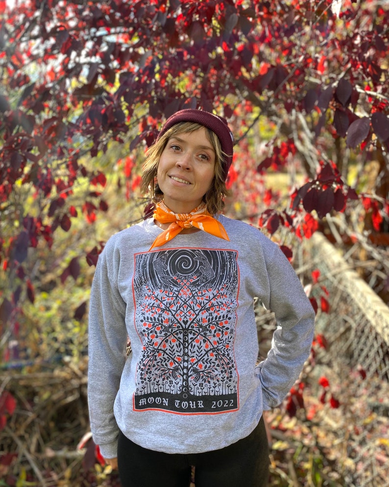 K&ouml;nnte beinhalten: Ein graues Sweatshirt mit einem schwarzen und orangenen Aufdruck eines Baumes mit V&ouml;geln und dem Text "MOON TOUR 2022". Die Person, die das Sweatshirt tr&auml;gt, steht vor einem Busch mit roten Bl&auml;ttern.