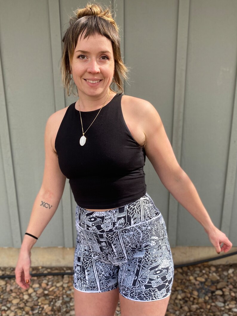 May include: A woman wearing a black tank top and black and white patterned high-waisted shorts. The shorts have a white outline and a variety of abstract designs.