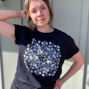 May include: A black t-shirt with a white floral design. The design is a circle with flowers and leaves arranged in a symmetrical pattern.