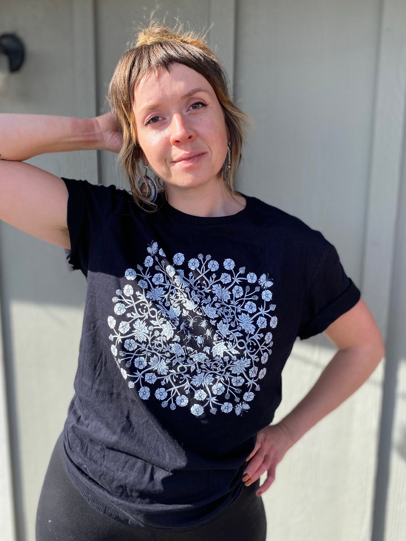 May include: A black t-shirt with a white floral design. The design is a circle with flowers and leaves arranged in a symmetrical pattern.