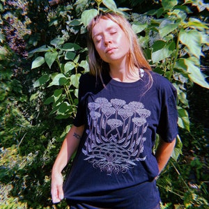 K&ouml;nnte beinhalten: Ein schwarzes T-Shirt mit einer wei&szlig;en Grafik einer stilisierten Pflanze mit mehreren Stielen und Bl&uuml;ten. Die Pflanze hat ein gro&szlig;es Wurzelsystem.