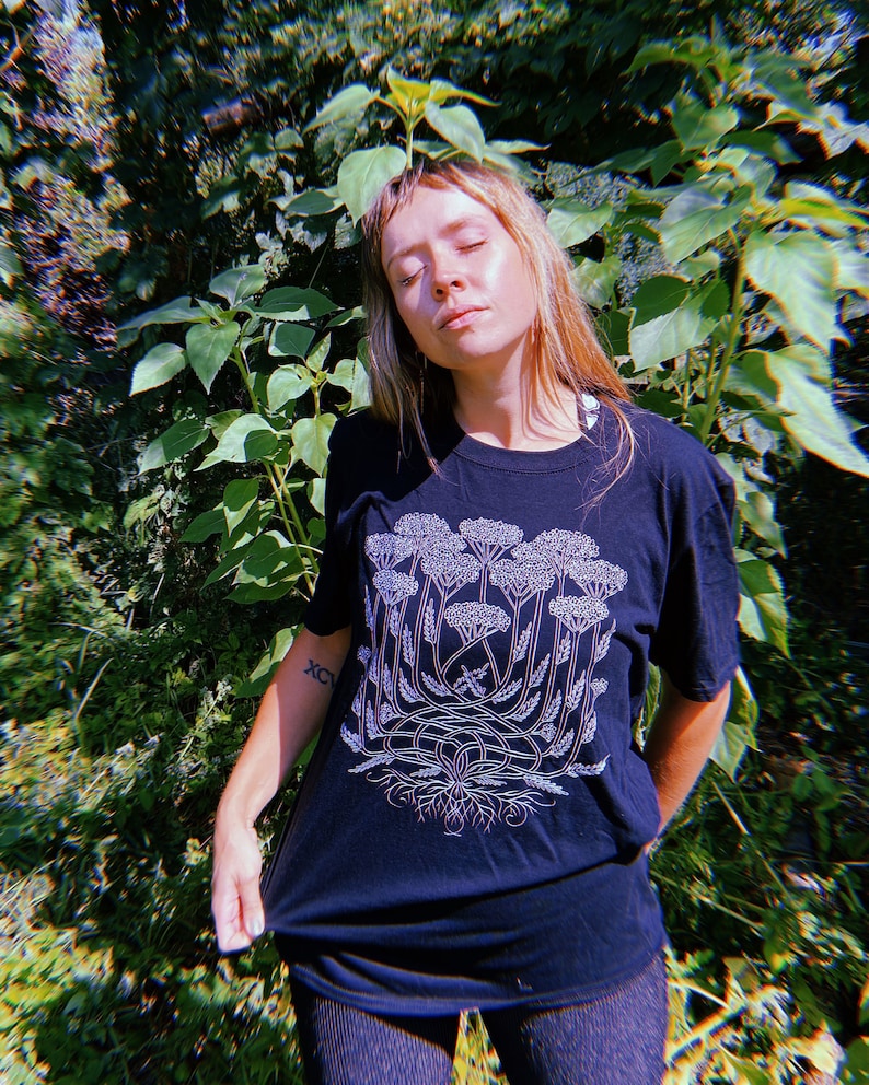 K&ouml;nnte beinhalten: Ein schwarzes T-Shirt mit einer wei&szlig;en Grafik einer stilisierten Pflanze mit mehreren Stielen und Bl&uuml;ten. Die Pflanze hat ein gro&szlig;es Wurzelsystem.
