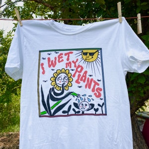 May include: White t-shirt with a graphic print of a cartoon flower watering a plant with the text "I Wet My Plants".