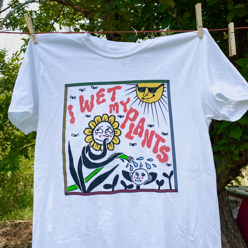 May include: White t-shirt with a graphic print of a cartoon flower watering a plant with the text "I Wet My Plants".