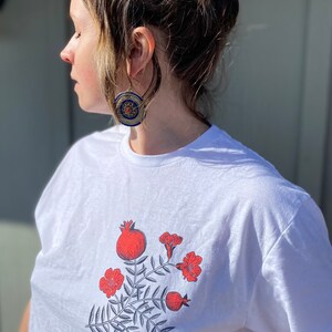 May include: White t-shirt with a graphic print of red pomegranates and flowers on a leafy branch.