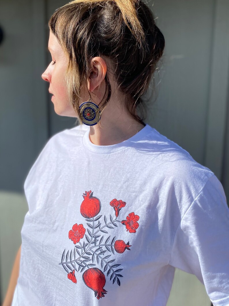 May include: White t-shirt with a graphic print of red pomegranates and flowers on a leafy branch.