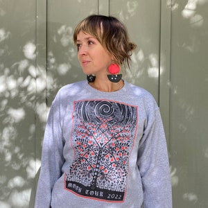 K&ouml;nnte beinhalten: Grauer Pullover mit einem schwarzen und roten grafischen Druck eines Baumes mit V&ouml;geln und dem Text "MOON TOUR 2022".