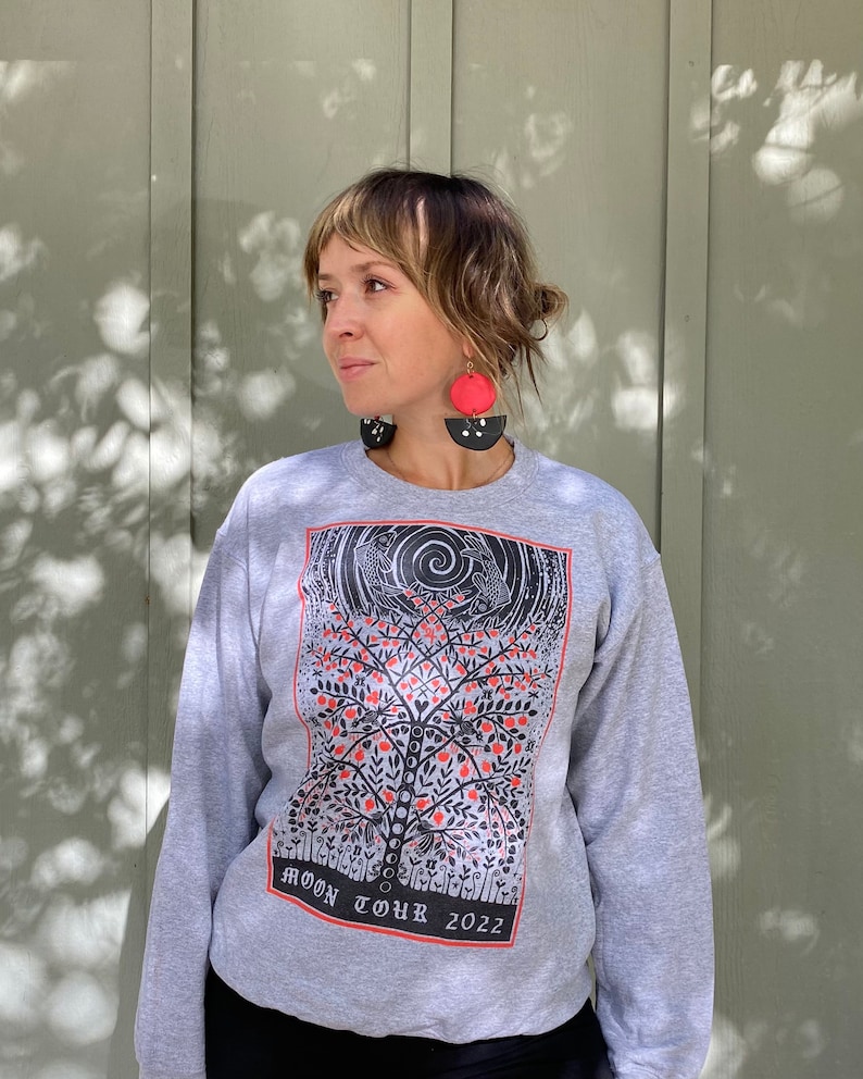 K&ouml;nnte beinhalten: Grauer Pullover mit einem schwarzen und roten grafischen Druck eines Baumes mit V&ouml;geln und dem Text "MOON TOUR 2022".