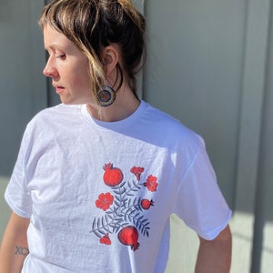 May include: White t-shirt with a red and blue floral design featuring pomegranates and leaves.