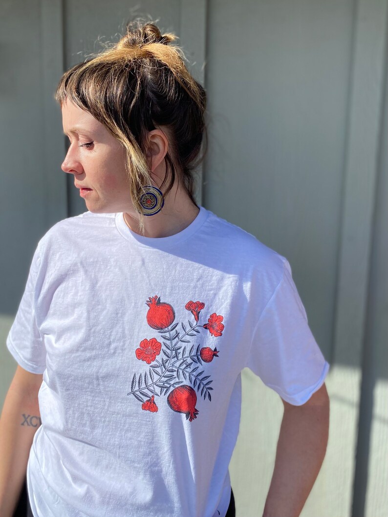 May include: White t-shirt with a red and blue floral design featuring pomegranates and leaves.