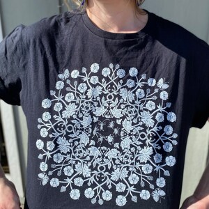 May include: Black t-shirt with a white floral design featuring a mandala pattern. The design includes small insects and butterflies.