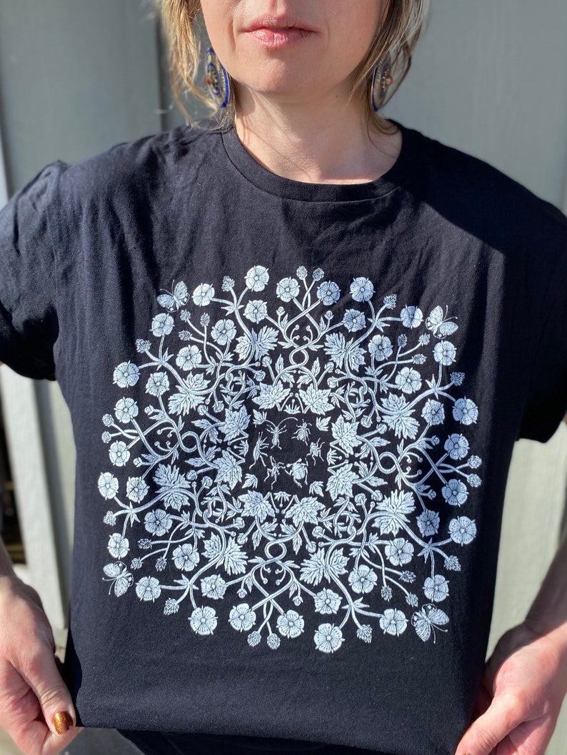 May include: Black t-shirt with a white floral design featuring a mandala pattern. The design includes small insects and butterflies.