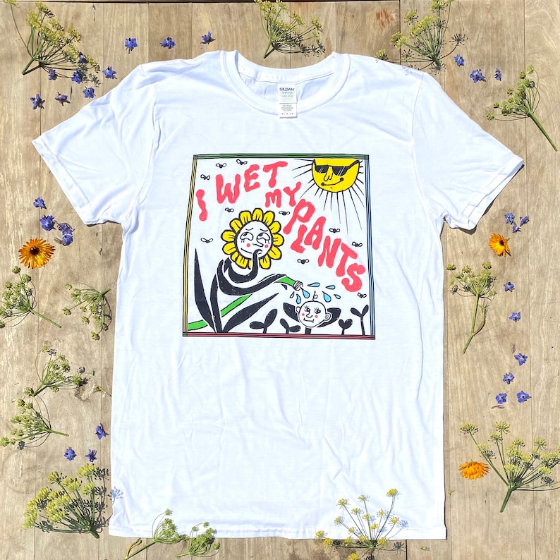 May include: White t-shirt with a cartoon illustration of a flower watering a plant with the text "I WET MY PLANTS" in a rainbow colored box.