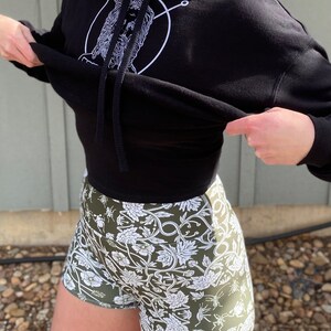 May include: Black cropped hoodie with a white graphic of a face inside a circle. The hoodie is paired with green athletic shorts with a white floral pattern.
