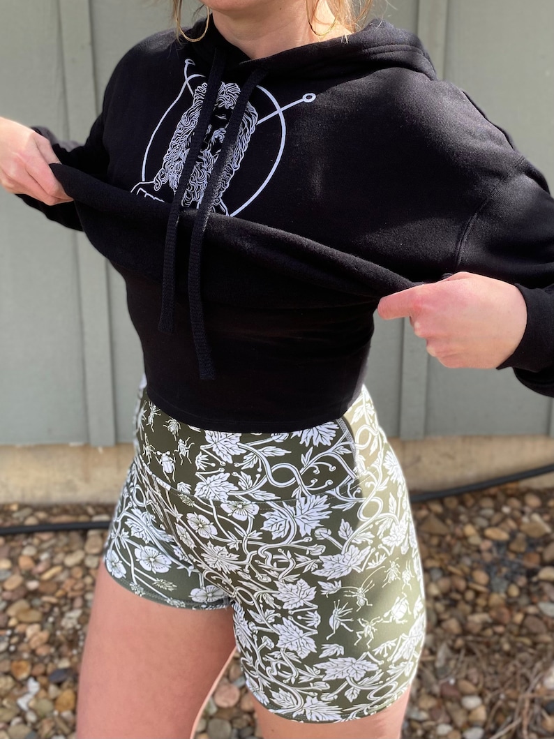 May include: Black cropped hoodie with a white graphic of a face inside a circle. The hoodie is paired with green athletic shorts with a white floral pattern.