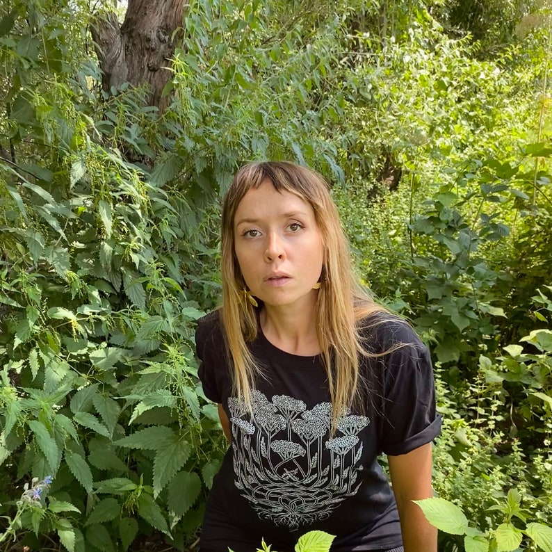 K&ouml;nnte beinhalten: Eine Frau mit langen blonden Haaren tr&auml;gt ein schwarzes T-Shirt mit einem wei&szlig;en Aufdruck einer Pflanze mit Blumen. Sie steht in einem Wald mit gr&uuml;nem Laub.