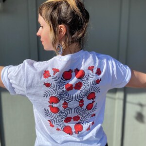 May include: White t-shirt with a red and black pomegranate design. The design features multiple pomegranates with leaves and flowers.