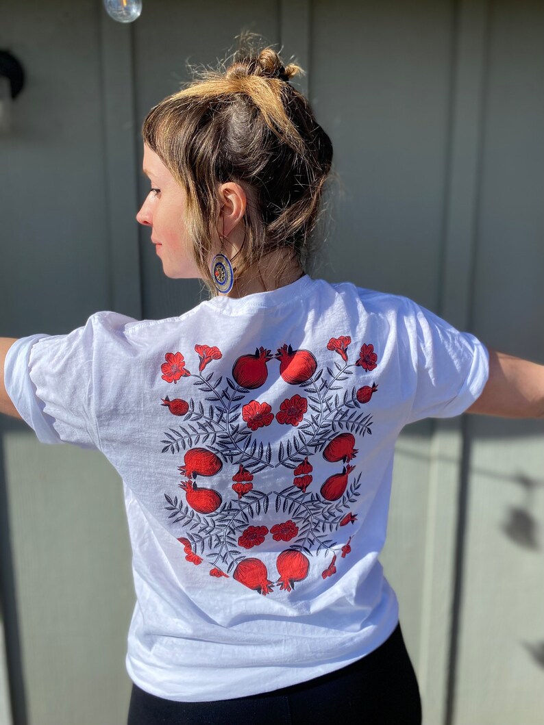 May include: White t-shirt with a red and black pomegranate design. The design features multiple pomegranates with leaves and flowers.