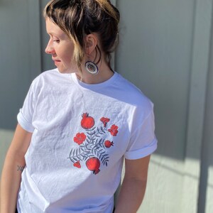May include: White t-shirt with a red and green floral design featuring pomegranates. The design is centered on the shirt.