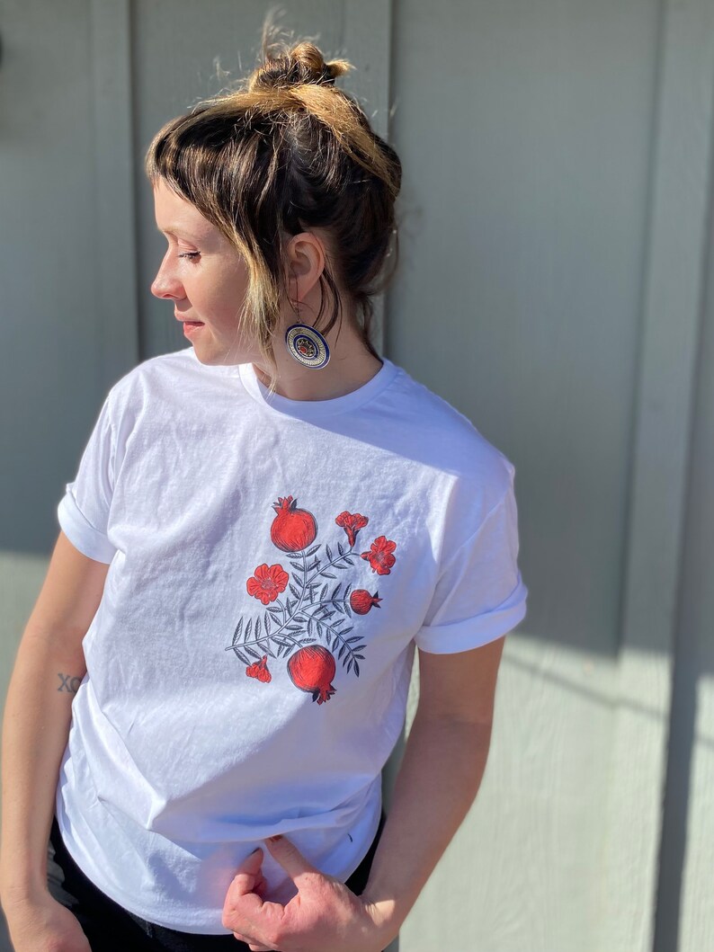May include: White t-shirt with a red and green floral design featuring pomegranates. The design is centered on the shirt.