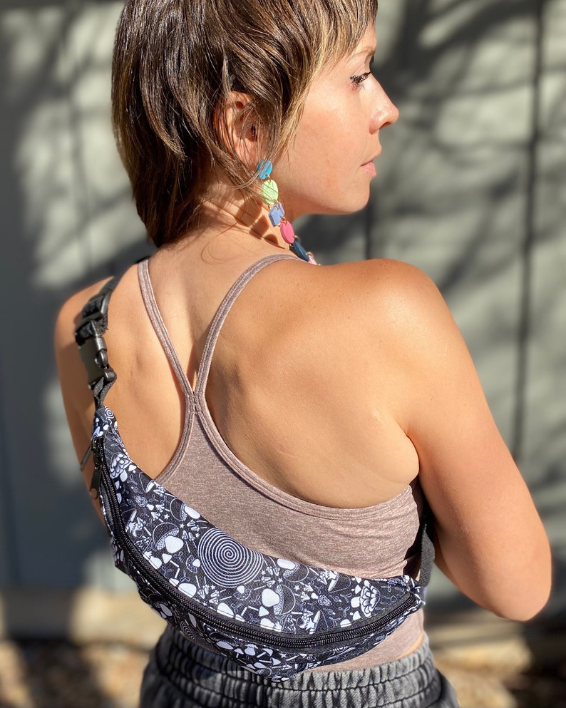 May include: A black fanny pack with a white and black psychedelic mushroom print. The fanny pack has a zipper closure and an adjustable strap.