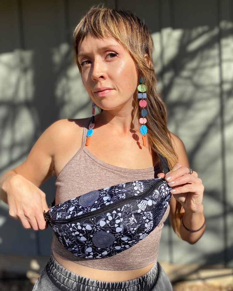 May include: A black fanny pack with a white psychedelic print featuring mushrooms, skulls, and other symbols. The fanny pack is worn by a person with long brown hair and colorful earrings.