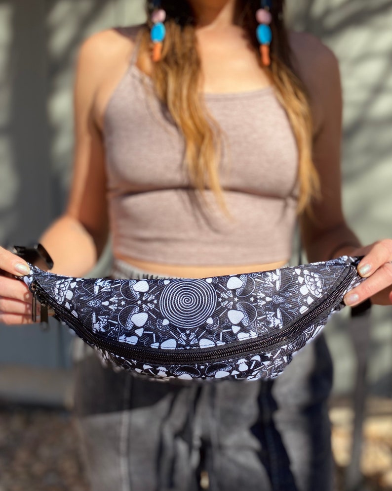 May include: A black fanny pack with a white psychedelic mushroom and spiral design. The fanny pack has a zipper closure.