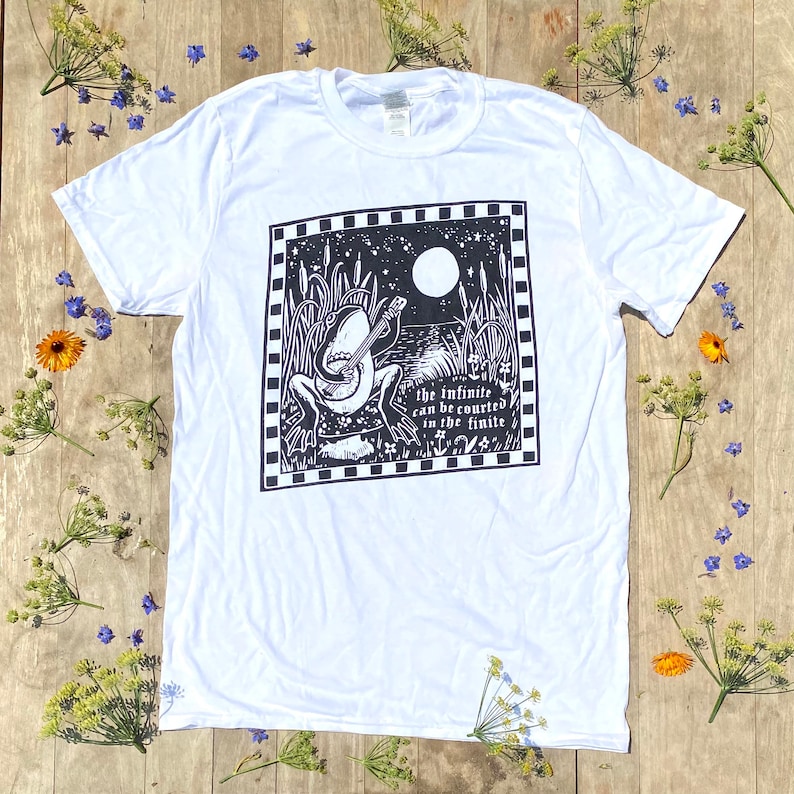 May include: White t-shirt with a black and white illustration of a frog playing a banjo. The illustration is surrounded by a black and white checkered border. The text "the infinite can be counted in the finite" is below the frog.