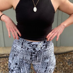 May include: Black tank top with a gold chain necklace and a white oval pendant. The person is wearing black and white patterned shorts with a graphic design.