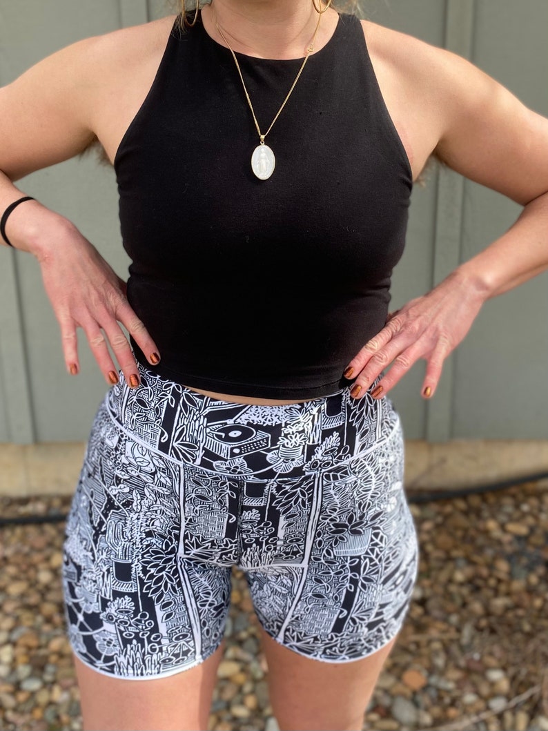 May include: Black tank top with a gold chain necklace and a white oval pendant. The person is wearing black and white patterned shorts with a graphic design.