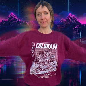 May include: A maroon sweatshirt with a graphic design featuring the word "COLORADO" in white block letters. The design includes a grid pattern and a stylized illustration of a figure. The text "NEON RIVALS" is below the graphic.
