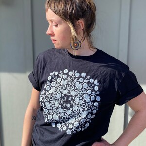 May include: A black t-shirt with a white floral mandala design. The mandala features a circular pattern of flowers and leaves.