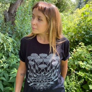 K&ouml;nnte beinhalten: Ein schwarzes T-Shirt mit einem wei&szlig;en Aufdruck einer Gruppe von Queen-Anne-Lace-Blumen. Der Aufdruck ist detailliert und zeigt die Stiele, Bl&auml;tter und Wurzeln der Blumen.