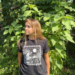 May include: A person wearing a black t-shirt with a white graphic of a person playing a guitar in a field with the text "The infinite can be counted in the hills".