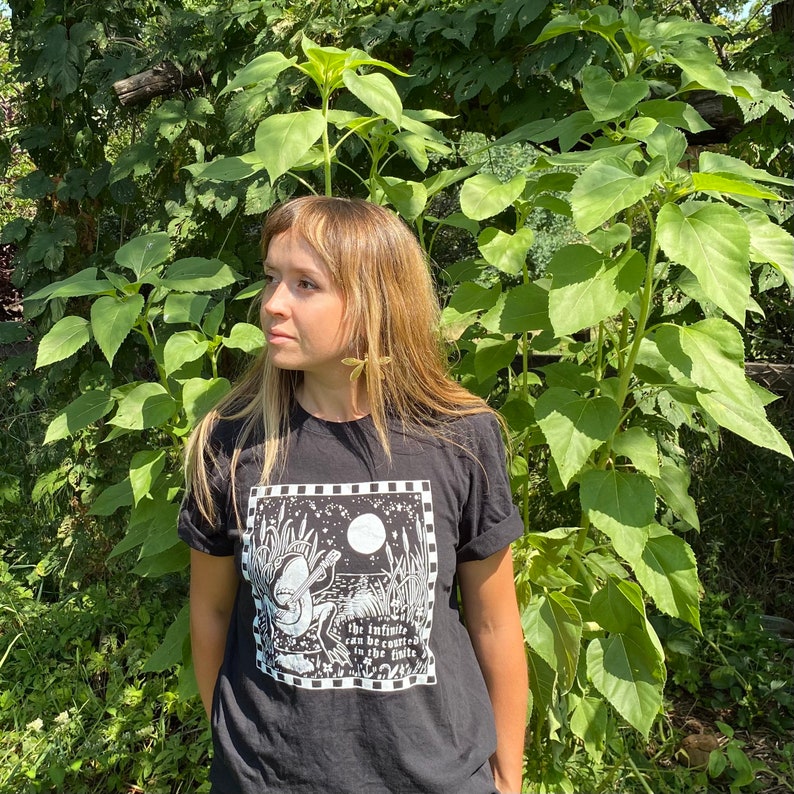 May include: A person wearing a black t-shirt with a white graphic of a person playing a guitar in a field with the text "The infinite can be counted in the hills".
