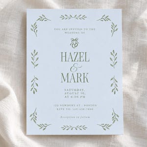 May include: A white rectangular wedding invitation card featuring a sage green botanical leaf border, a small illustration of two rings, and text reading Hazel & Mark, Saturday, August 10, at 6:00 PM, 123 Newbury St., Boston, RSVP to 123-456-7890.
